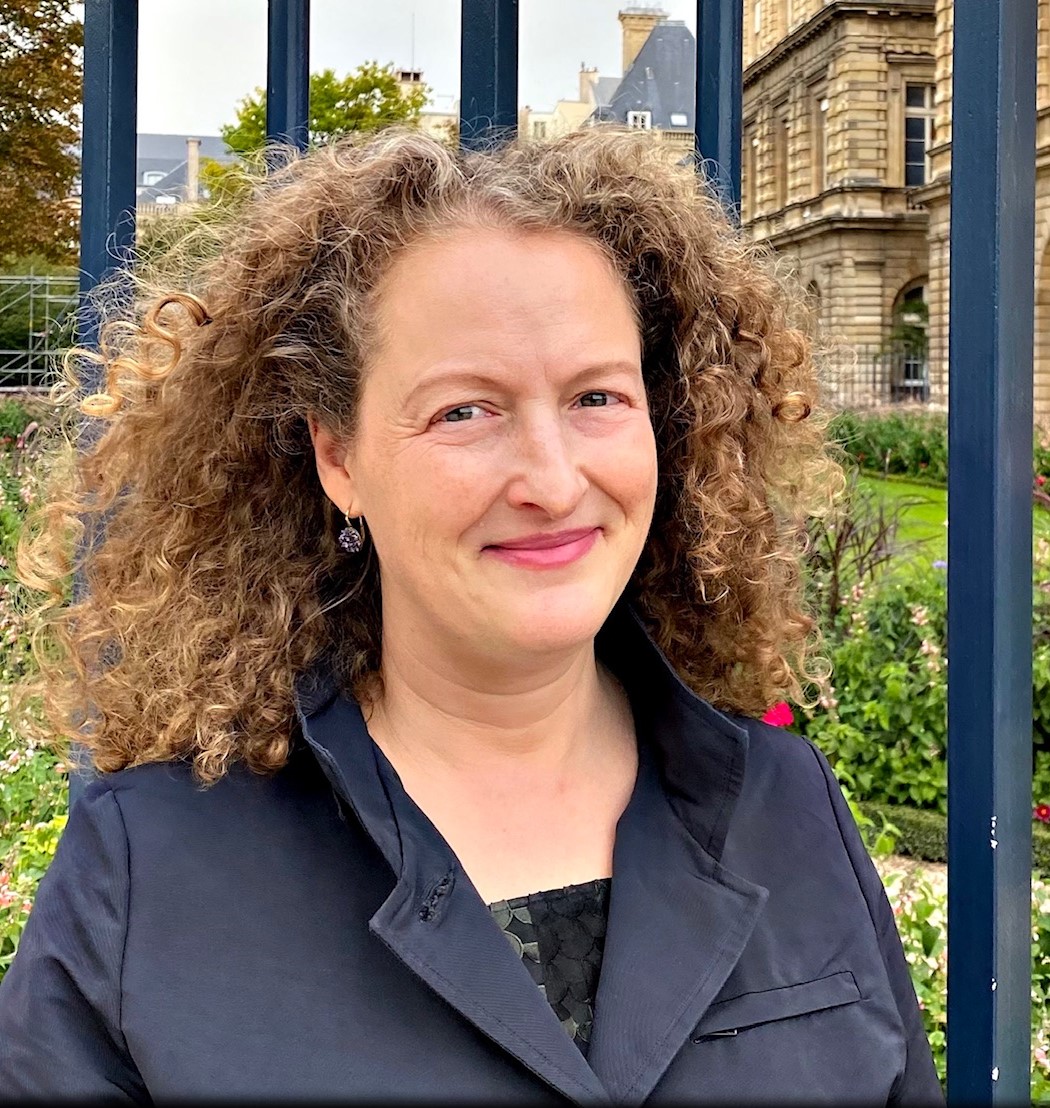Asha Agrawal has curly light brown hair and stands in front of a historical european style looking building
