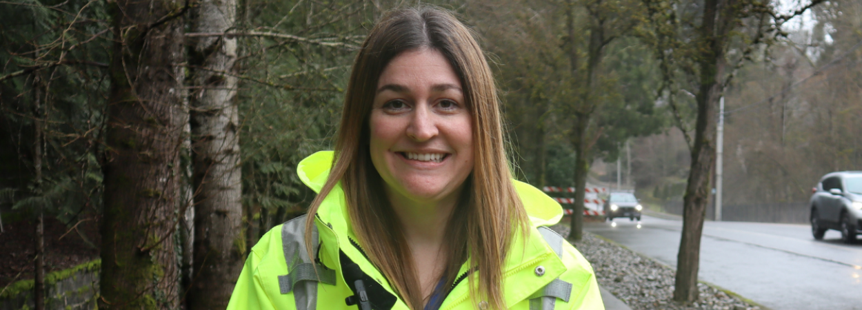 Courtney Furman stands on the sidewalk next to a wooded area on one side and a road with cars passing. She has long brown hair and a highlighter yellow rain jacket. 