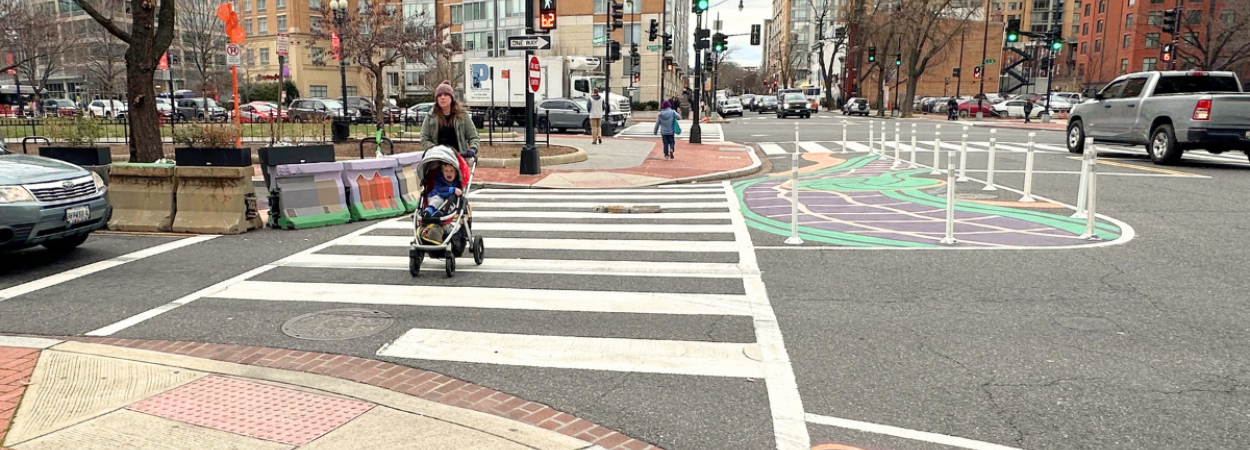A person pushing a baby stroller with a young child inside crosses an intersection with a tactical curb extension with mural art installation.