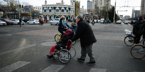 A woman on a yellow bicycle and a person pushing another person in a wheelchair through a crosswalk