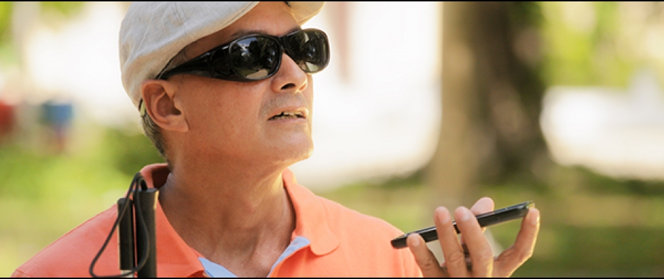 A man with sunglasses and a cane carries a smartphone in his hand, appearing to be listening to its audio