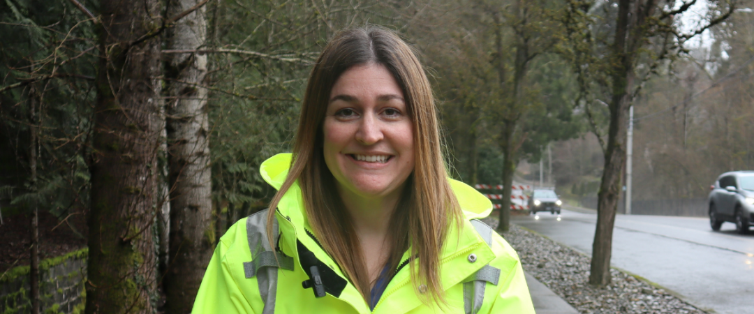 Courtney Furman stands on the sidewalk next to a wooded area on one side and a road with cars passing. She has long brown hair and a highlighter yellow rain jacket. 