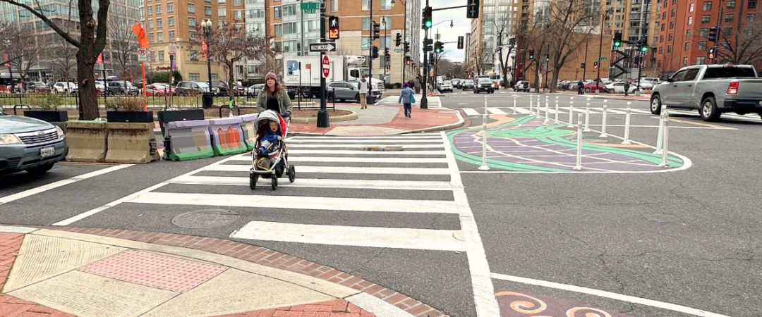A person pushing a baby stroller with a young child inside crosses an intersection with a tactical curb extension with mural art installation.